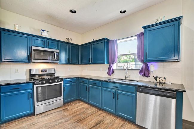 Bright kitchen with striking blue cabinets and stainless steel appliances. Features a gas stove, dishwasher, and microwave. Natural light comes through a window above the sink. Wood-look flooring adds warmth. | Image 22