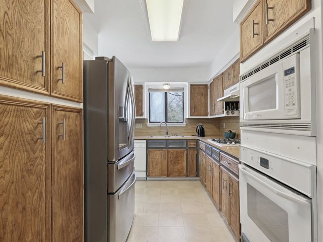 Kitchen with white appliances, backsplash, under cabinet range hood, brown cabinetry, and a sink | Image 8