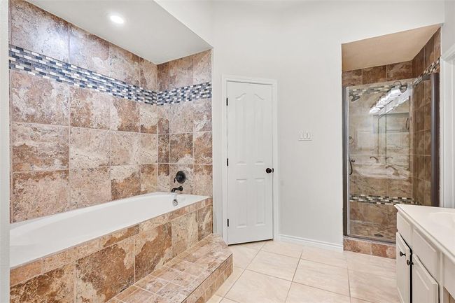 The primary bathroom features separate vanities with updated lighting, dual walk-in closets, a deep soaking tub with decorative tile surround and step-up entry, plus a spacious walk-in shower accented by coordinating mosaic tile detailing. | Image 18