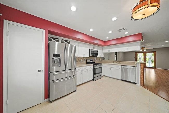 This modern kitchen features stainless steel appliances, white cabinetry, and a stylish red accent wall. It opens into a spacious living area with wood vinyl flooring and ample natural light. | Image 13