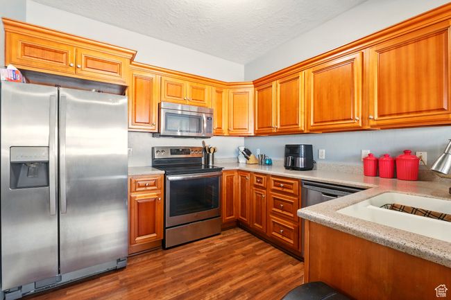 Kitchen with appliances with stainless steel finishes, dark wood finished floors, brown cabinetry, a textured ceiling, and a sink | Image 12