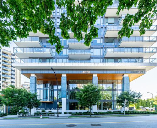 Modern building exterior featuring extensive glass paneling, multiple balconies with glass railings, a spacious ground-level entrance with large windows, and a landscaped perimeter with mature trees | Card Image