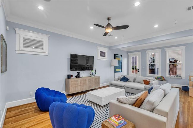 Bright and inviting family room with light grey walls, recessed lighting, and a ceiling fan. Hardwood floors and large windows enhance natural light. | Image 12