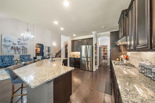 The kitchen showcases a modern kitchen with dark wood cabinetry, granite countertops, and stainless steel appliances. The space opens to a living area with elegant seating and a chandelier, creating a welcoming atmosphere. A walk-in pantry is visible, adding ample storage. | Image 9