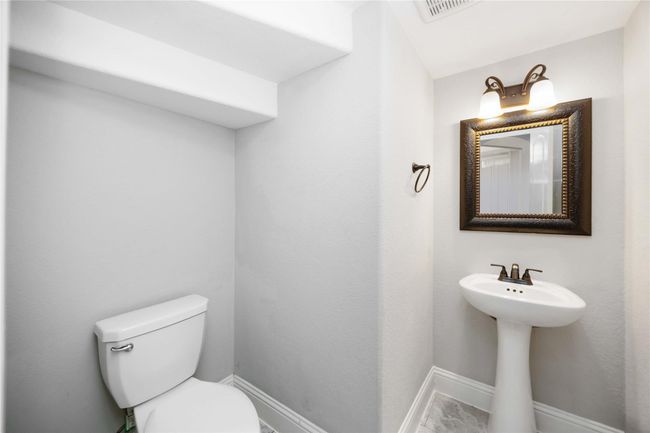 This is a compact, modern half-bathroom featuring a pedestal sink, a toilet, and a decorative framed mirror. The walls are painted in a light neutral color, and the lighting fixture adds a warm touch. | Image 8