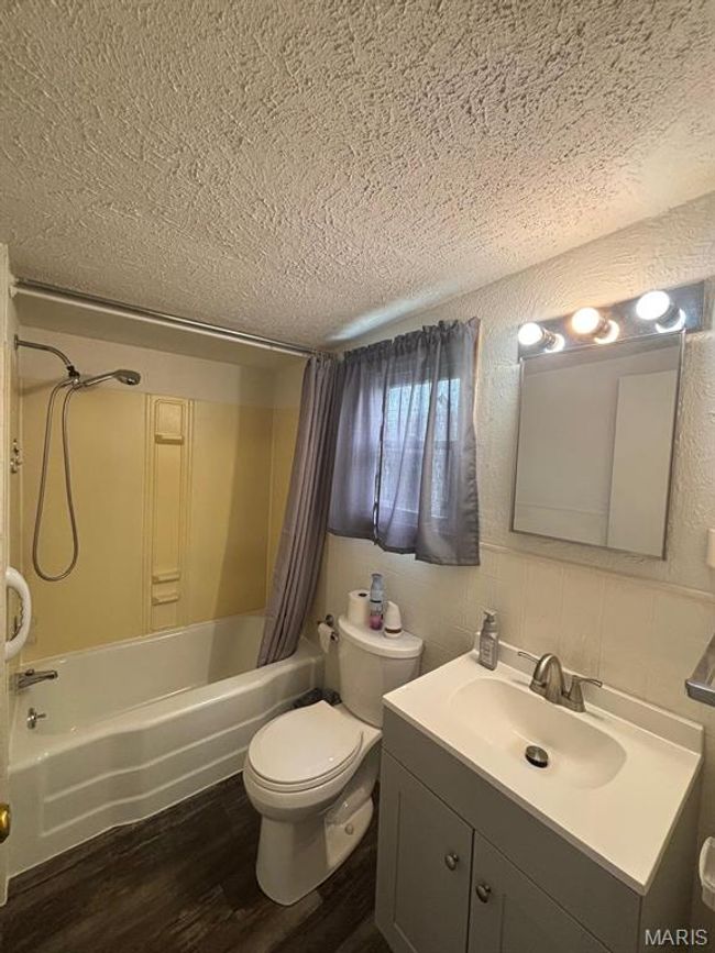 Full bath featuring a textured ceiling, vanity, shower / bathtub combination with curtain, wood finished floors, and a textured wall | Image 22