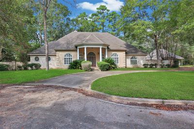 This is a single-story home with a tan stucco exterior, arch detailing around the front door, and a shingle roof. It features a curved driveway, mature trees, and well-kept landscaping, suggesting a serene and private setting. | Image 3