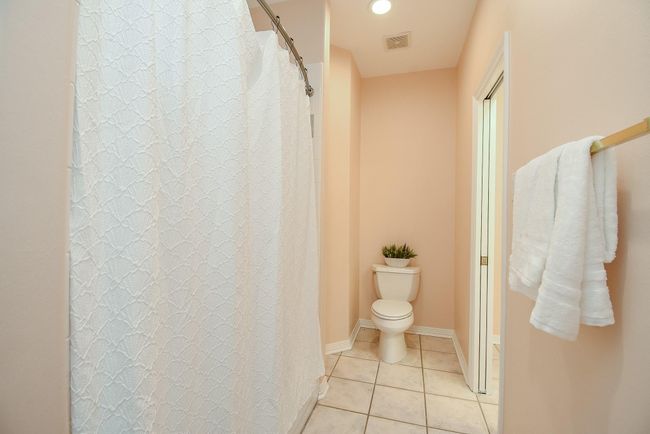 This bathroom features soft peach walls, a tiled floor, a shower with a white curtain, and a neatly placed towel on a rack. A toilet area is enhanced by a small plant for a touch of greenery. | Image 39