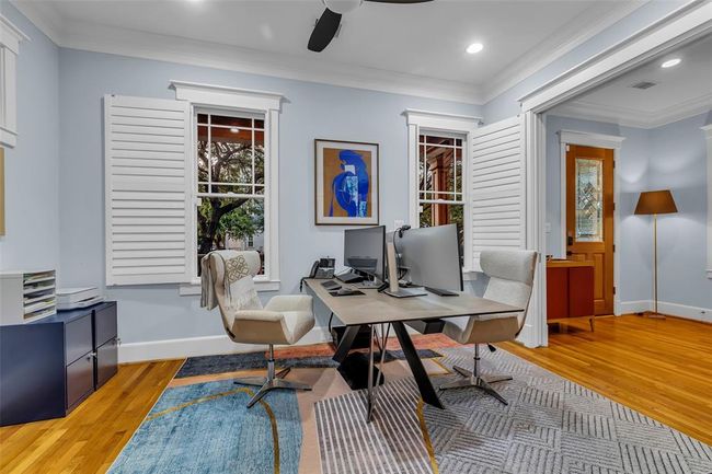 This spacious and bright home office features hardwood floors, two large windows with shutters, and a stylish desk setup. The space provides a comfortable and functional work environment. | Image 9