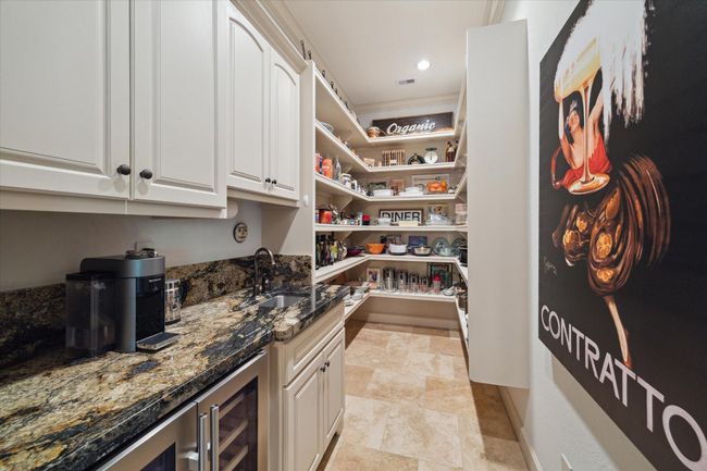 This spacious walk-in pantry goes beyond storage, featuring extensive built-in shelving for organized, easy access to dry goods and serving pieces. A dedicated prep area includes exotic granite countertops, upper soft-close cabinetry, a dual beverage refrigerator, and a built-in sink—perfect for a coffee bar, cocktail setup, or catering support. Tucked just off the kitchen, this space blends practicality with elegance for everyday ease or effortless entertaining. | Image 19