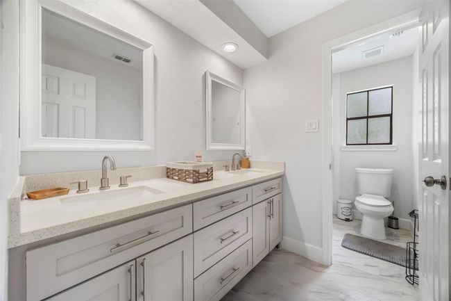 A bathroom featuring a double-sink vanity with sleek fixtures, ample storage, and a large mirror. The space extends to a separate toilet area with a frosted window for natural light, complemented by neutral tones and elegant tile flooring. | Image 15