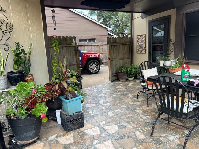 Charming patio area with decorative tile flooring, surrounded by potted plants. Features outdoor seating and a view of a wooden gate leading to the driveway. Cozy and inviting for relaxation or entertaining. | Image 11
