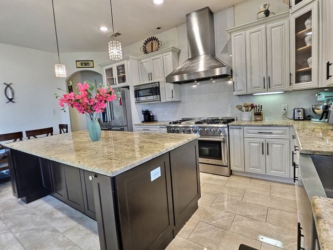 Kitchen featuring wall chimney range hood, appliances with stainless steel finishes, a center island, light stone countertops, and hanging light fixtures | Image 12
