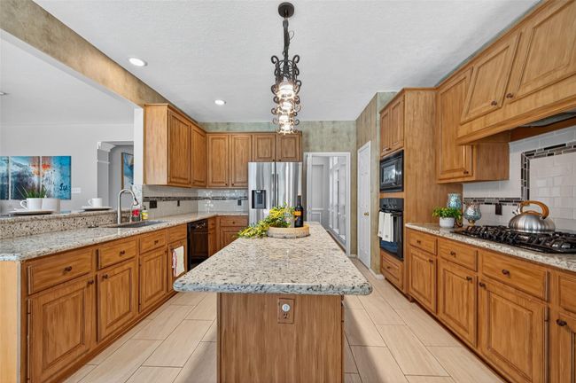 This spacious kitchen features warm wooden cabinetry, granite countertops, and modern stainless steel appliances, including a gas stove. The central island provides ample workspace and seating, while stylish pendant lighting adds a touch of elegance. | Image 12