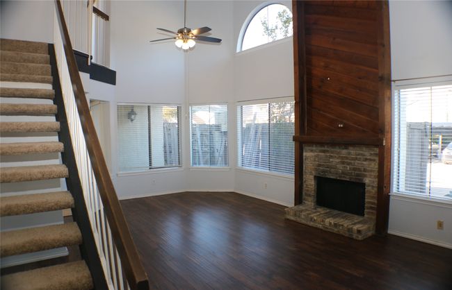 Unfurnished living room featuring a ceiling fan, wood finished floors, stairs, a brick fireplace, and a high ceiling | Image 9