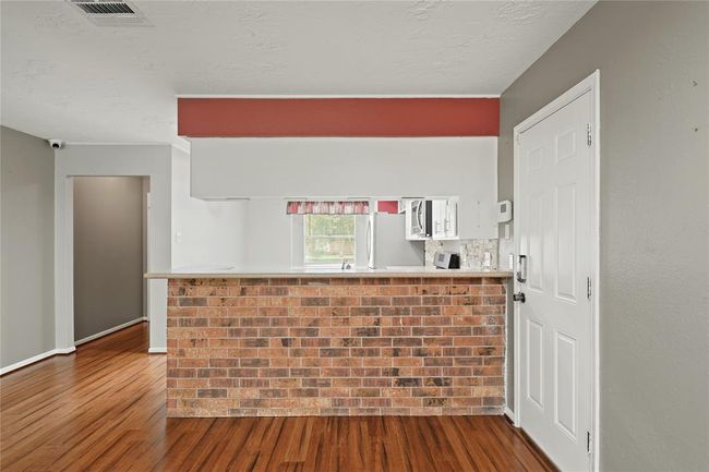 This photo shows a view of the cozy kitchen area through a brick accented counter opening from the living area. The kitchen is partially visible, featuring stainless steel appliances and a window with a view. The layout is open and inviting. | Image 6
