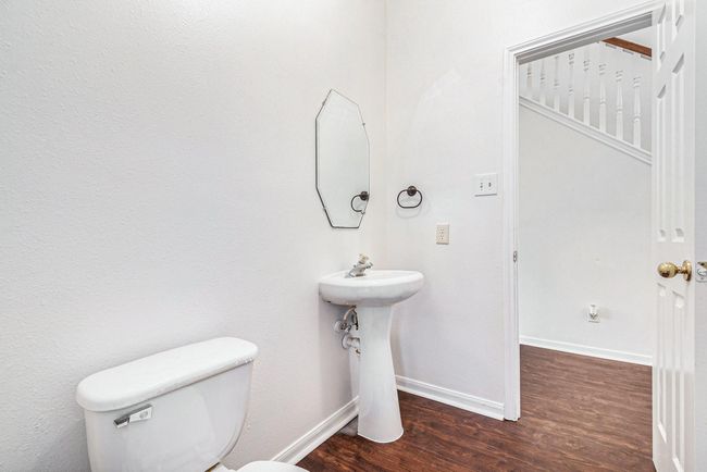 This photo showcases a small, clean half-bathroom with white walls and a simple pedestal sink. It features a wall-mounted mirror, a toilet, and dark wood flooring. The space is adjacent to office and the home's main hallway. | Image 8