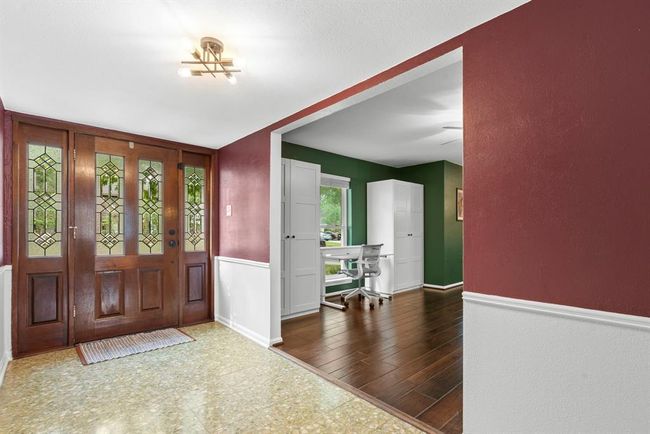 This photo showcases a welcoming entryway with a stylish wood and glass door, leading to a cozy formal living room featuring rich green walls and warm wooden flooring. | Image 5