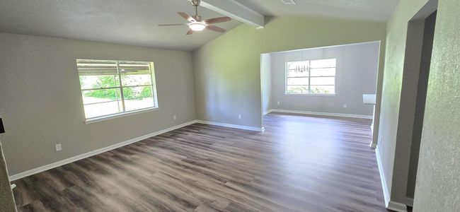 This photo shows a spacious, open living area with modern luxury vinyl plank flooring and neutral walls. It features a vaulted ceiling and seamlessly flows into the dining area with another window and a central light fixture, creating a bright and inviting space. | Image 9