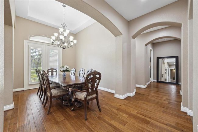 This elegant dining area set under a stylish chandelier. The room is brightened by large windows with shutters, and it opens to a hallway with arched doorways and warm wood flooring, creating a welcoming and sophisticated atmosphere. | Image 4