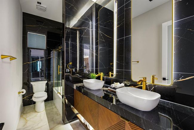 This striking secondary bathroom features bold black marble-style walls with gold veining, elevated by modern gold fixtures and dual vessel sinks. The large, illuminated mirrors and sleek wood vanity add a luxurious touch, while a glass-enclosed tub and shower combo offers both style and functionality. | Image 30