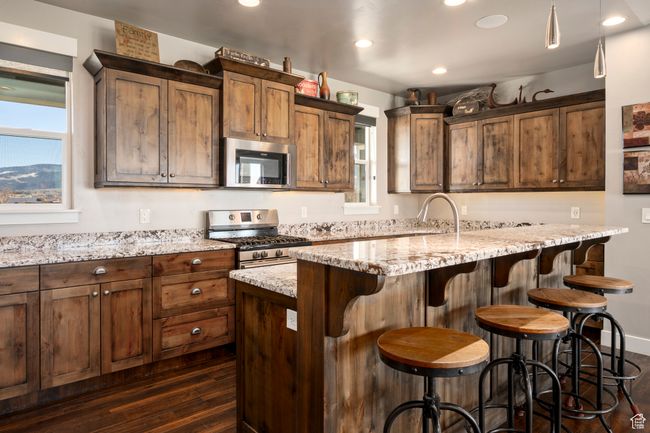 Kitchen with dark wood-style flooring, a breakfast bar, an island with sink, light stone countertops, and stainless steel appliances | Image 11