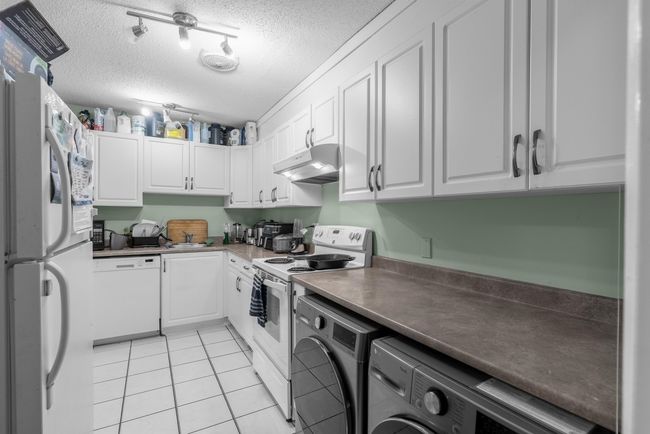 Kitchen with white appliances, a textured ceiling, light tile patterned flooring, white cabinets, and under cabinet range hood | Image 8