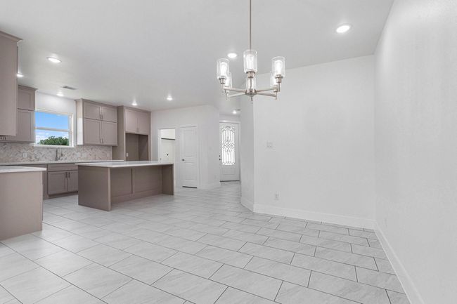 This photo showcases a spacious, modern kitchen and dining area. It features sleek cabinetry, a large island, light tile flooring, and a contemporary chandelier. The area is well-lit with recessed lighting and natural light from a window. | Image 9