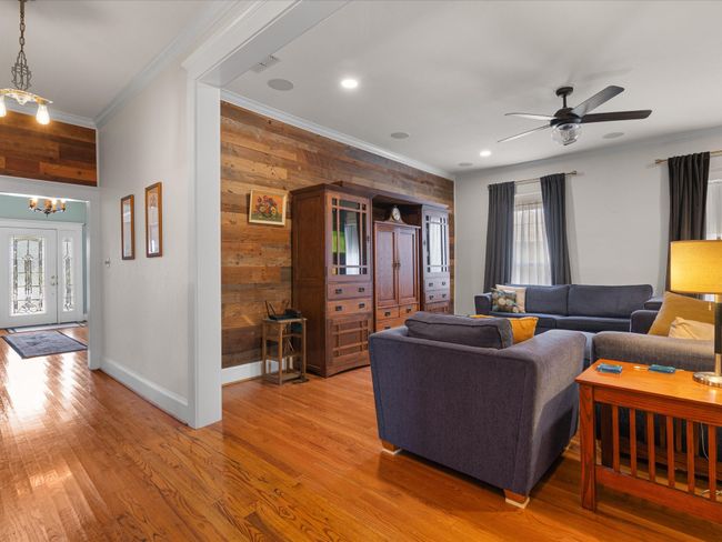 Living area featuring light wood-type flooring, ornamental molding, ceiling fan with notable chandelier, wooden walls, and baseboards | Image 11