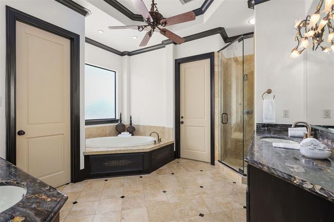 This luxurious bathroom features a spacious layout with a corner soaking tub, elegant black marble countertops, and a glass-enclosed shower. The room is accented by a ceiling fan, stylish lighting, and large frosted window for natural light. | Image 35