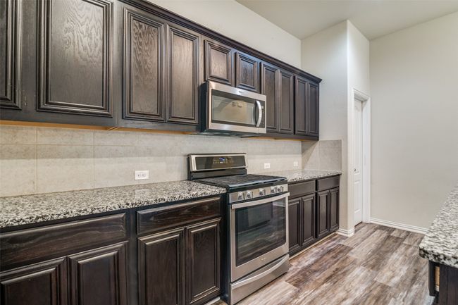 Kitchen featuring appliances with stainless steel finishes, wood finished floors, decorative backsplash, dark brown cabinetry, and light stone countertops | Image 12