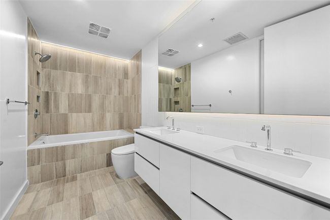 The primary bathroom showcases a dual vanity with sleek white cabinets, providing ample storage & a modern aesthetic plus a large horizontal mirror, enhancing the spacious feel and functionality of the area. The shower and tub combo is designed with floor to ceiling tiles. | Image 9