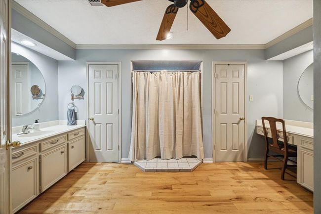 Primary ensuite bathroom features dual sinks with large mirrors, ample cabinet storage, and a central shower area. It has hardwood flooring, a ceiling fan, and a vanity area with a chair, offering a blend of functionality and comfort. | Image 16