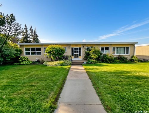 Exterior featuring light brick siding, white-framed windows, a concrete walkway, mature trees, and landscaped shrubbery | Card Image