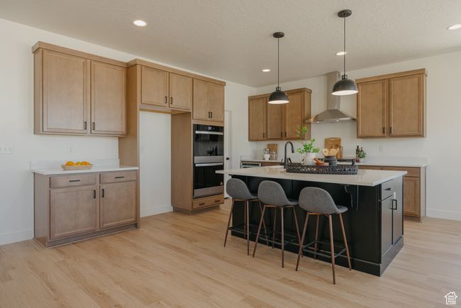 Kitchen with wall chimney range hood, stainless steel double oven, a kitchen island with sink, and light wood-type flooring | Image 4