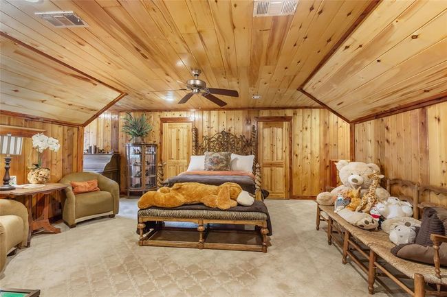 This cozy bedroom features warm wood paneling on walls and ceiling, creating a rustic cabin feel. It includes a comfortable bed, plush seating area, and ample space for relaxation. The room is adorned with charming decorations and a ceiling fan for comfort. | Image 42