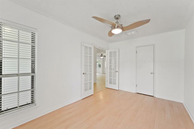 Bright room with light wood flooring, white walls, and a modern ceiling fan. Features a large window with blinds, French doors leading to another space, and a simple closet door. | Image 7
