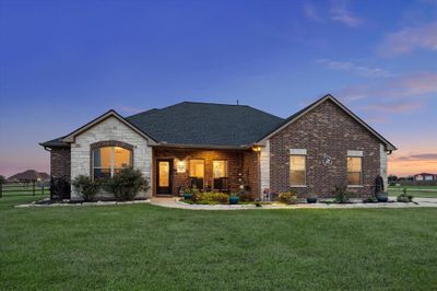 This is a single-story brick home featuring a well-manicured lawn and an inviting entrance with a warm, lit porch. The property enjoys an expansive, open sky backdrop, perfect for enjoying evening sunsets. | Image 2