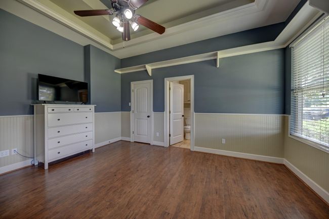 Guest room features walls with wainscoting. It includes a large window for natural light, a doorway to an adjacent bathroom, and a decorative tray ceiling. | Image 22