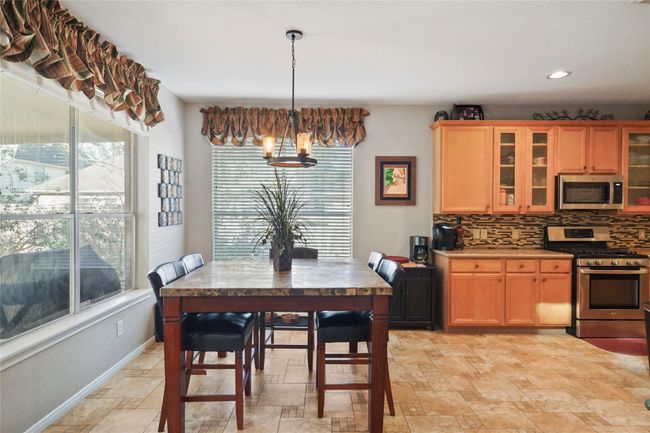 Bright kitchen with tile flooring, featuring a dining area. Large windows with decorative valances provide ample natural light. The kitchen includes wooden cabinets, a modern tile backsplash, and stainless steel appliances. | Image 10