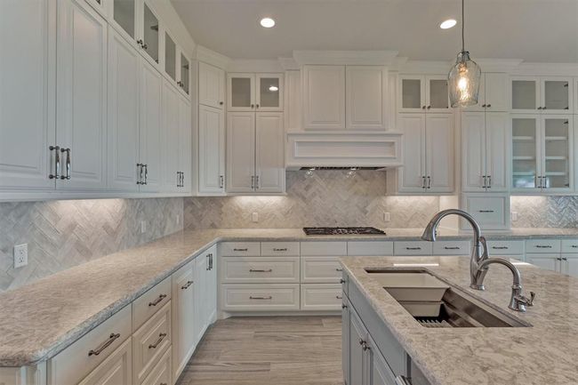 A high-end Thermador gas cooktop is paired with a custom vent hood, combining performance with designer appeal. The 42" upper cabinets, accented with glass display cabinets, add visual height and elegance. A striking herringbone-patterned backsplash brings just the right amount of texture and dimension, elevating the kitchen’s overall aesthetic. | Image 16