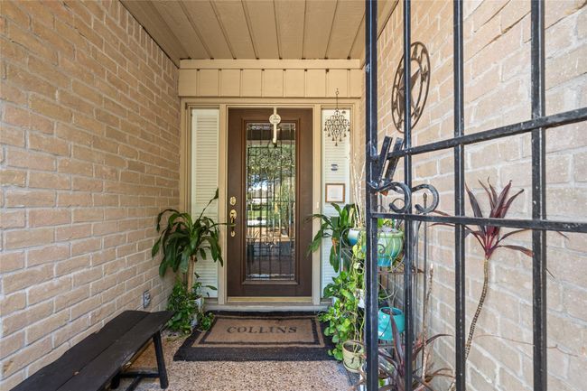 The welcoming front entryway features a charming brick exterior and a decorative iron gate for added security. A glass-paneled door allows natural light to filter in, creating a warm and inviting atmosphere. Lush greenery and potted plants enhance the cozy ambiance, while the covered porch provides a shaded retreat. | Image 6