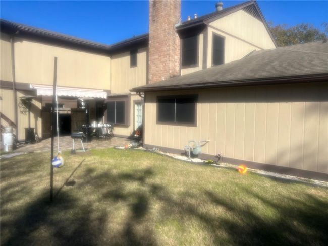 This photo shows a backyard with a grassy area and a patio. There's an outdoor seating arrangement and a barbecue grill. The house exterior is beige with a brick chimney, and there's a shade awning over the patio. | Image 15