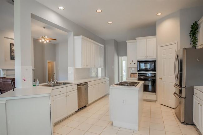 This kitchen features a spacious, open layout with white cabinetry, and a center island with a cooktop. It has tiled flooring and ample natural light, creating a bright and inviting space. | Image 23