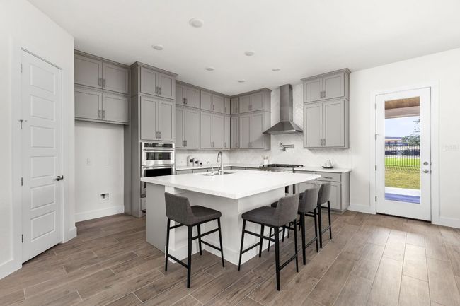 Kitchen with wall chimney exhaust hood, gray cabinets, wood finished floors, decorative backsplash, and a kitchen bar | Image 13