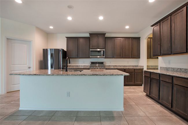 Kitchen featuring stainless steel appliances, dark brown cabinets, light stone countertops, a kitchen island with sink, and arched walkways | Image 13