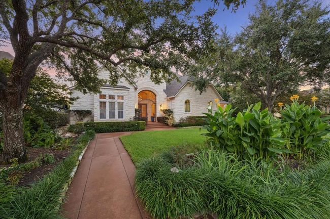 This charming home features a beautiful stone exterior with large arched windows and a welcoming front entry. Surrounded by lush greenery and mature trees, the pathway leads through well-maintained gardens, creating an inviting and tranquil atmosphere. | Image 4