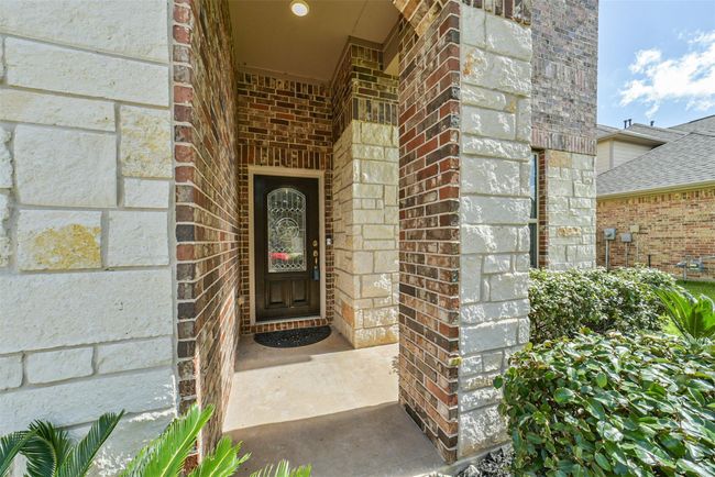Charming entrance with a covered porch featuring a mix of stone and brick. The welcoming front door has an elegant glass insert, and the area is landscaped with lush greenery. | Image 33