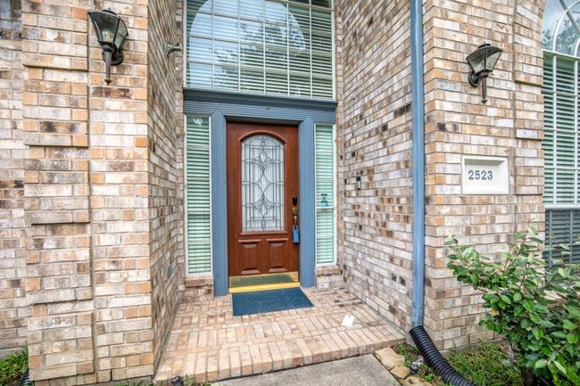 The entrance of a home with a welcoming arched wooden door featuring decorative glass panels, framed by light brickwork. Flanking the door are large windows, and there are wall-mounted lights on either side. | Image 7