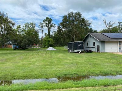 The photo shows a single-story home with a gray exterior and a detached garage, featuring solar panels on the roof. The property includes a well-maintained lawn with scattered mature trees. A puddle of water suggests recent rain in a tranquil, green setting. | Image 3